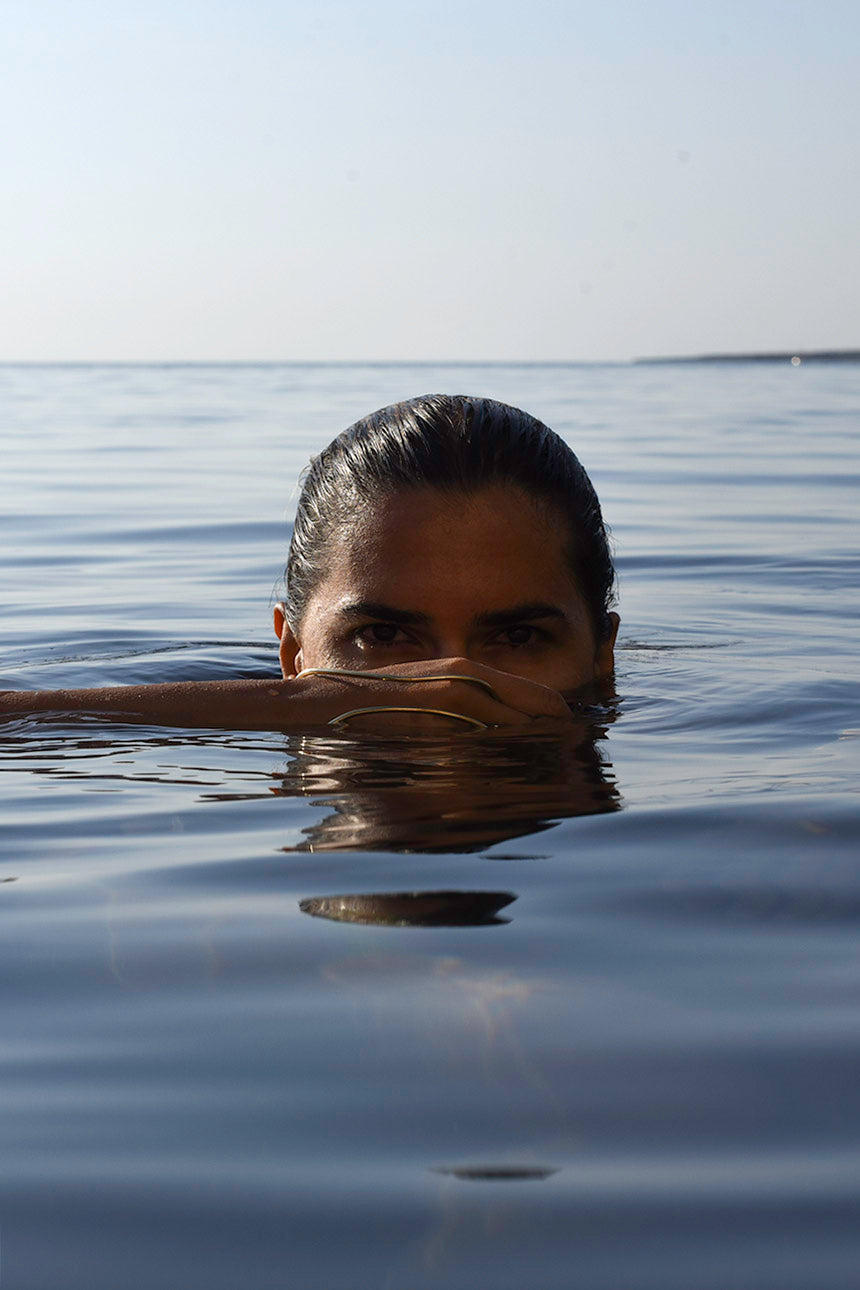 A woman with dark hair pulled back sits in calm blue water, only her eyes and upper face visible above the surface. Her hand showing the Delos Handmade Hand Bracelet made by MVDT.