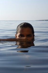 A woman with dark hair pulled back sits in calm blue water, only her eyes and upper face visible above the surface. Her hand showing the Delos Handmade Hand Bracelet made by MVDT.