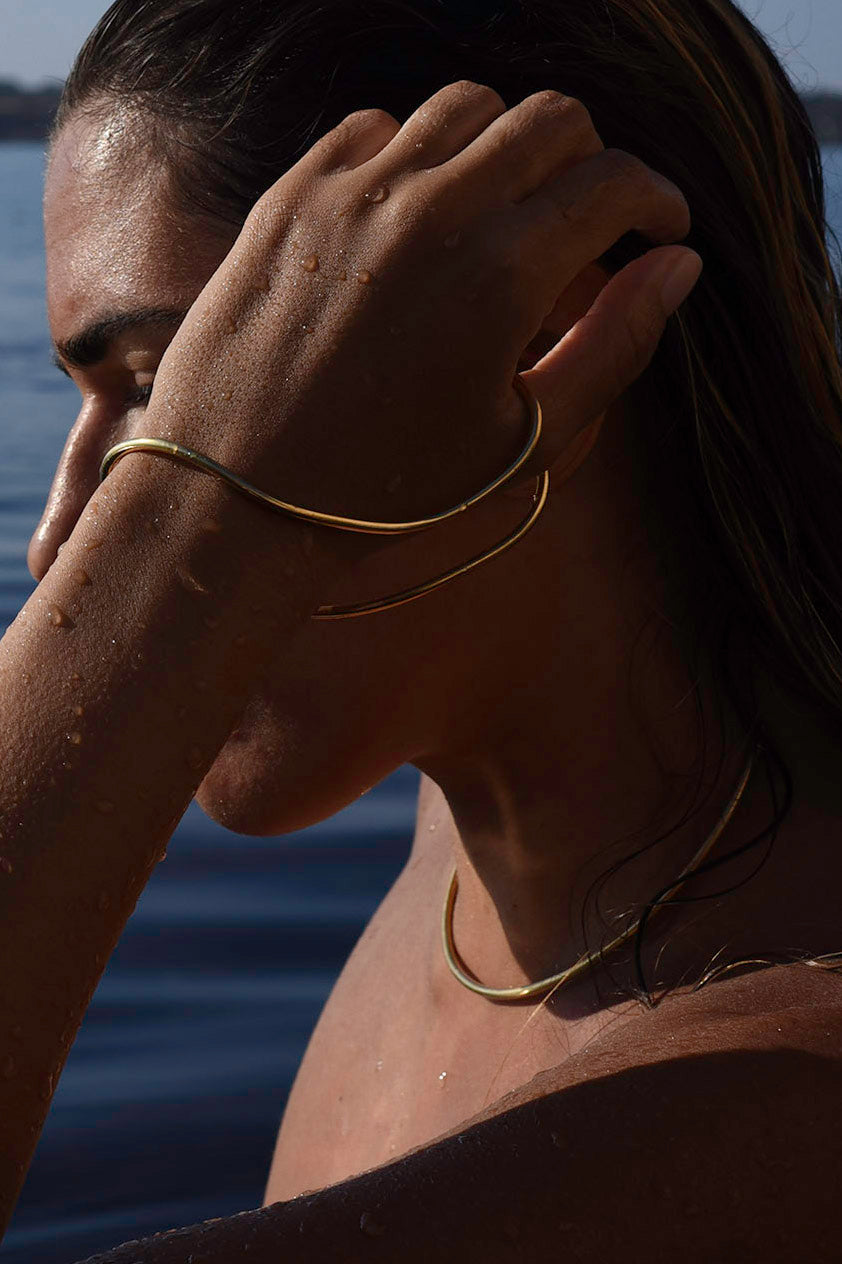 A close-up lifestyle shot of a woman with wet skin wearing minimalist gold jewelry, including a thin gold bangle, thumb-hand bracelet on her wrist and a delicate gold choker necklace, with water and a blurred outdoor background.