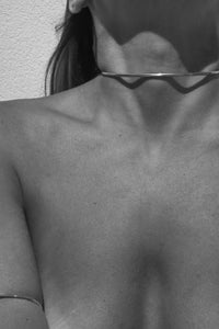 A close-up black and white photo of a person's bare shoulders and upper chest wearing a very thin, wavy choker necklace and a simple bangle on the upper arm. Handcrafted choker and arm cuff in gold made out of brass