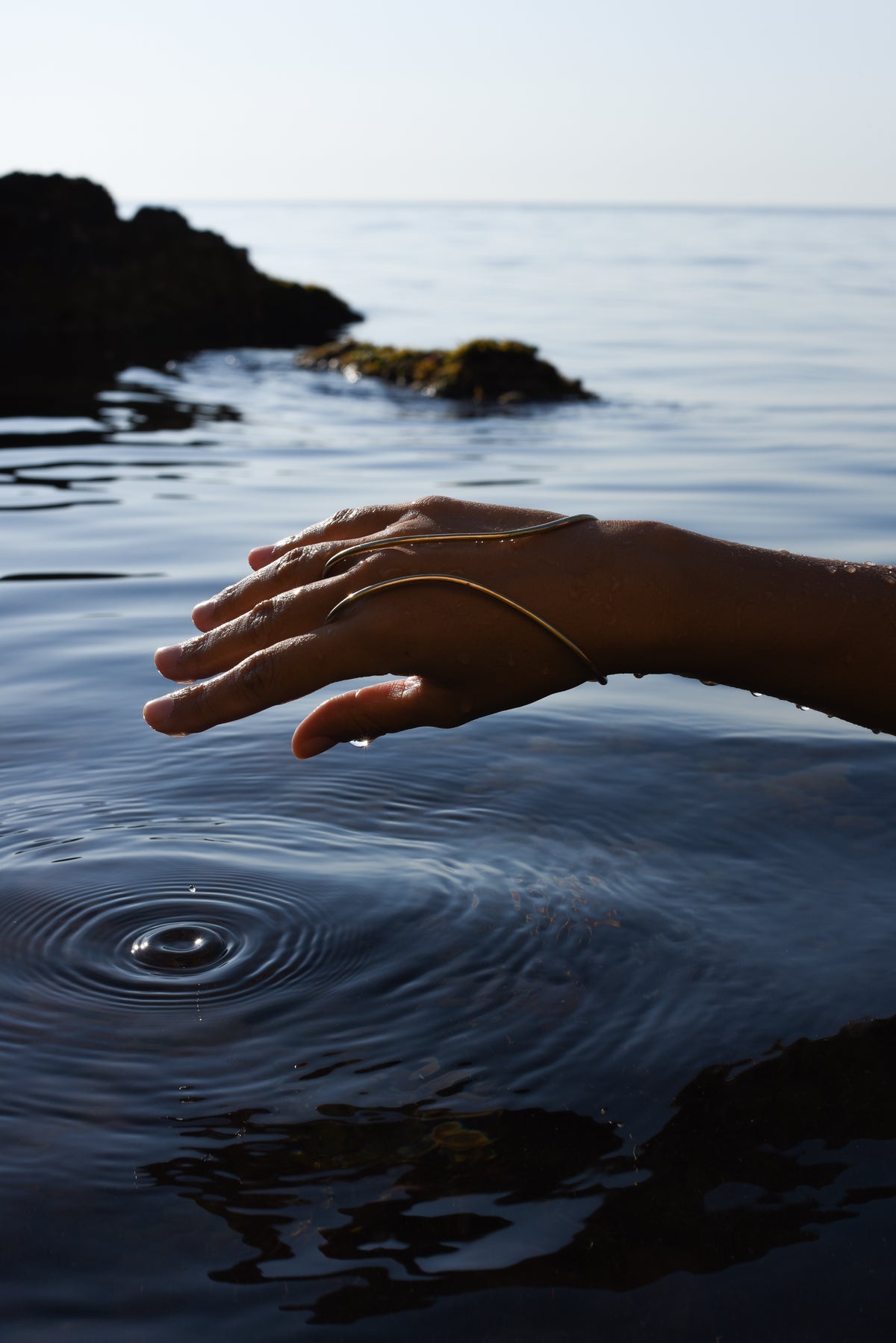 A person's hand and wrist wearing a delicate handcrafted gold hand bracelet connected as gold ring, resting near the surface of calm ocean water that creates ripples.