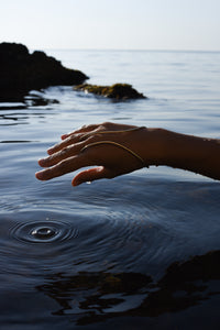 A person's hand and wrist wearing a delicate handcrafted gold hand bracelet connected as gold ring, resting near the surface of calm ocean water that creates ripples.