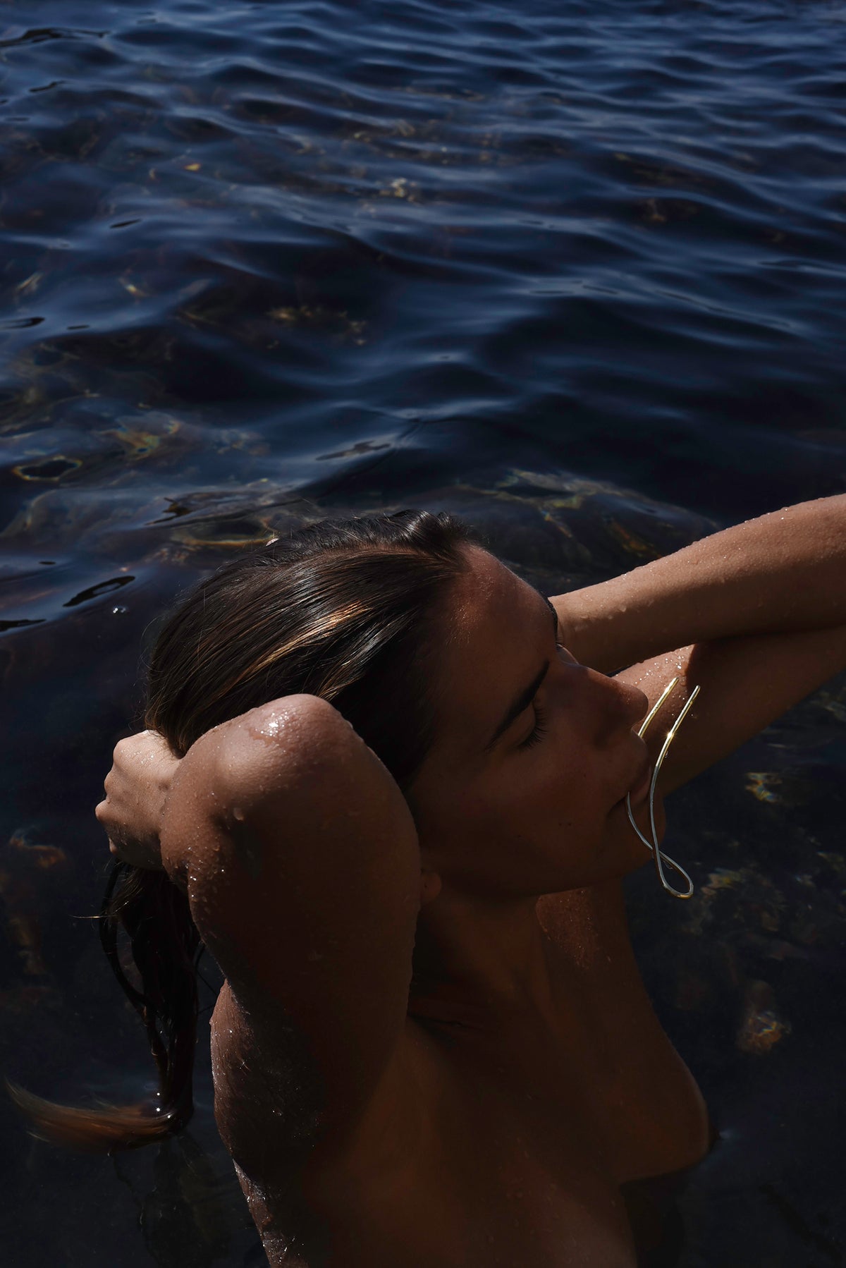 Woman coming out from the Mediterranean Sea holding the gold Delos hair pin in her mouth while making a knot of her hair, MVDT Collection Ortigia.