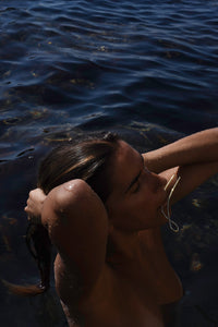 Woman coming out from the Mediterranean Sea holding the gold Delos hair pin in her mouth while making a knot of her hair, MVDT Collection Ortigia.