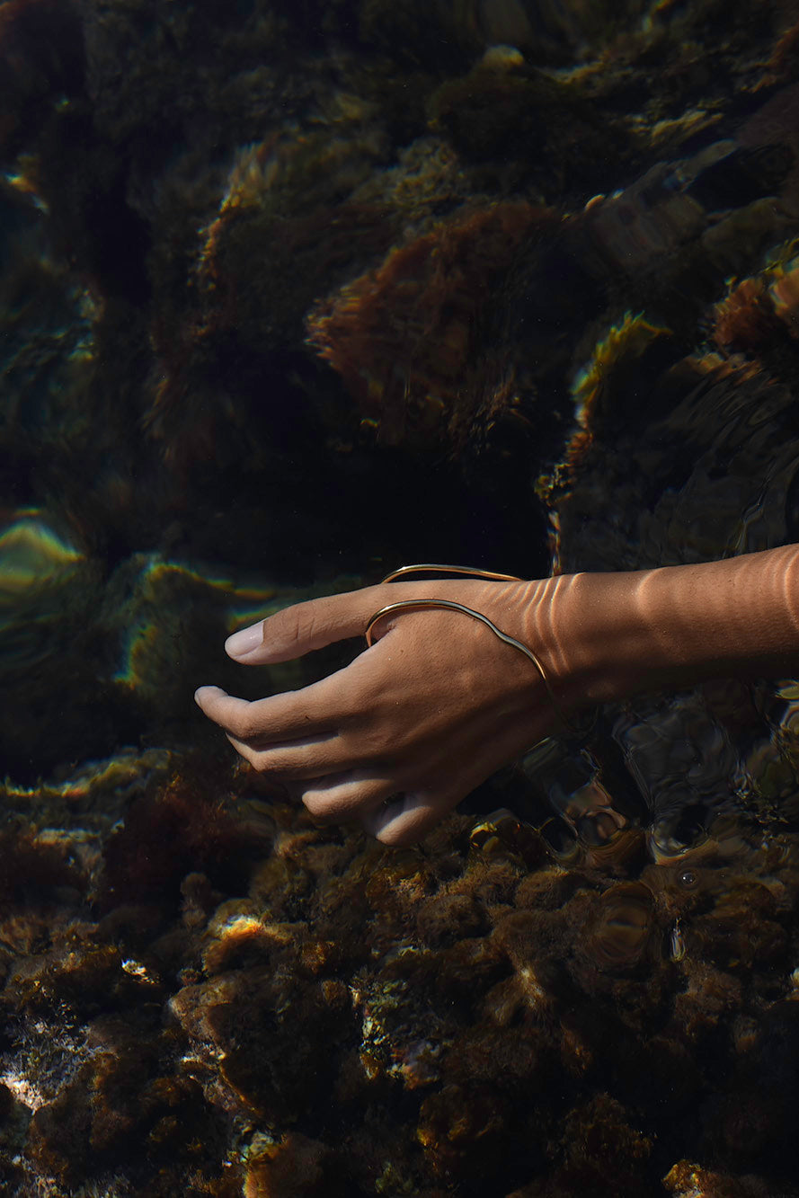A person's hand and wrist wearing a thin gold snake-like bangle, set against a dark, underwater rocky background with shallow water ripples. It's a handcrafted thumb-hand-bracelet.